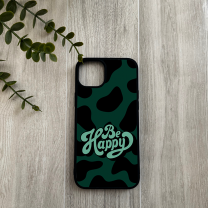 Phone case with 'Be Happy' text on a camouflage pattern, placed on a wooden surface with a leafy branch.