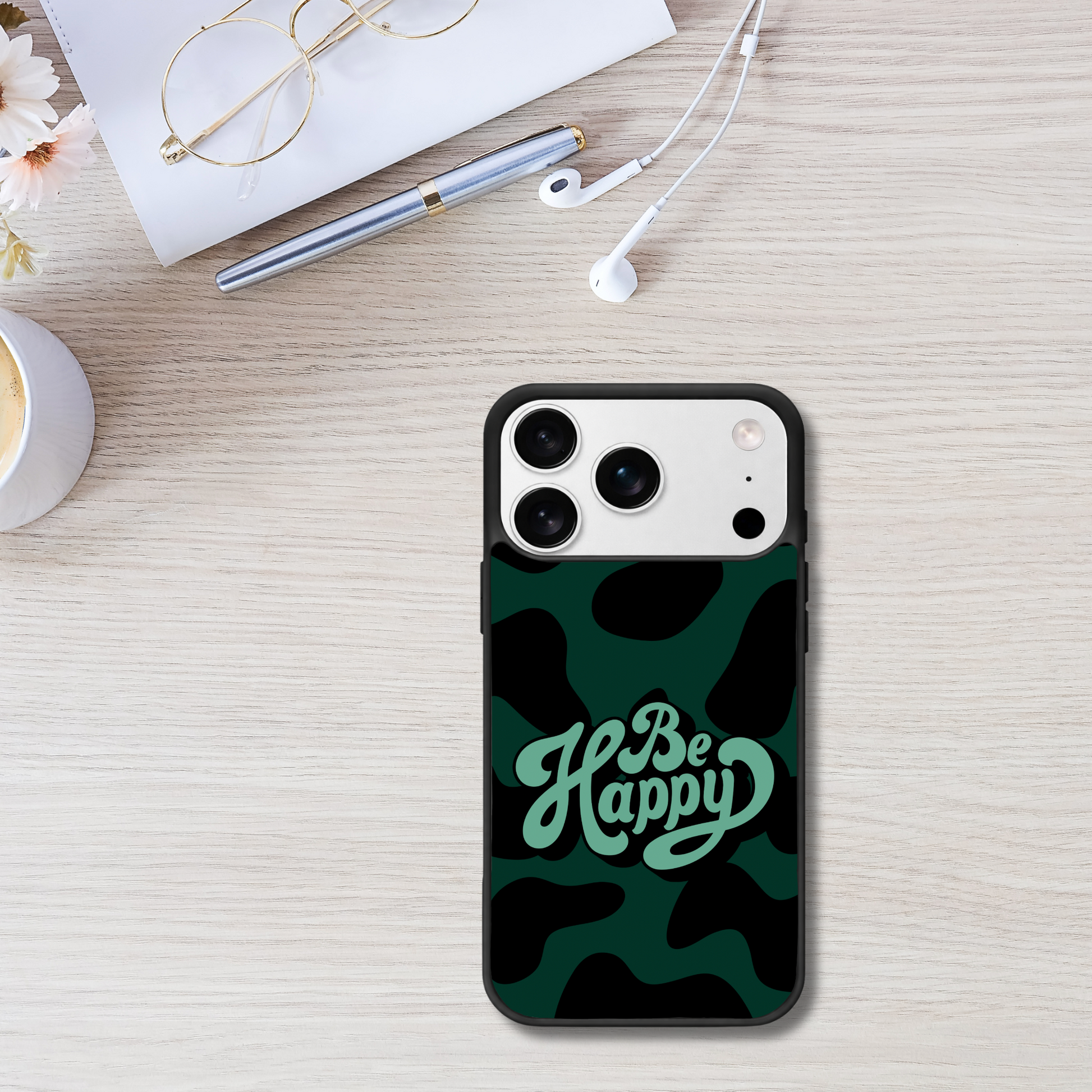 Phone case with 'Be Happy' text on a camouflage pattern, placed on a desk with glasses, pen, and flowers.