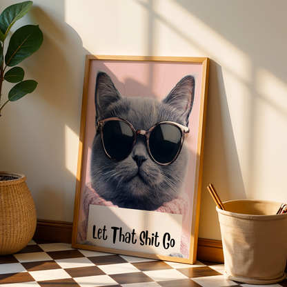 Framed picture of a cat wearing sunglasses with text 'Let That Shit Go' on a wall next to a plant and a small bucket.