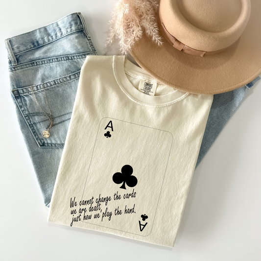 Ace of Clubs graphic t-shirt with motivational quote – "We cannot change the cards we are dealt, just how we play the hand" – laid flat with jeans and a hat